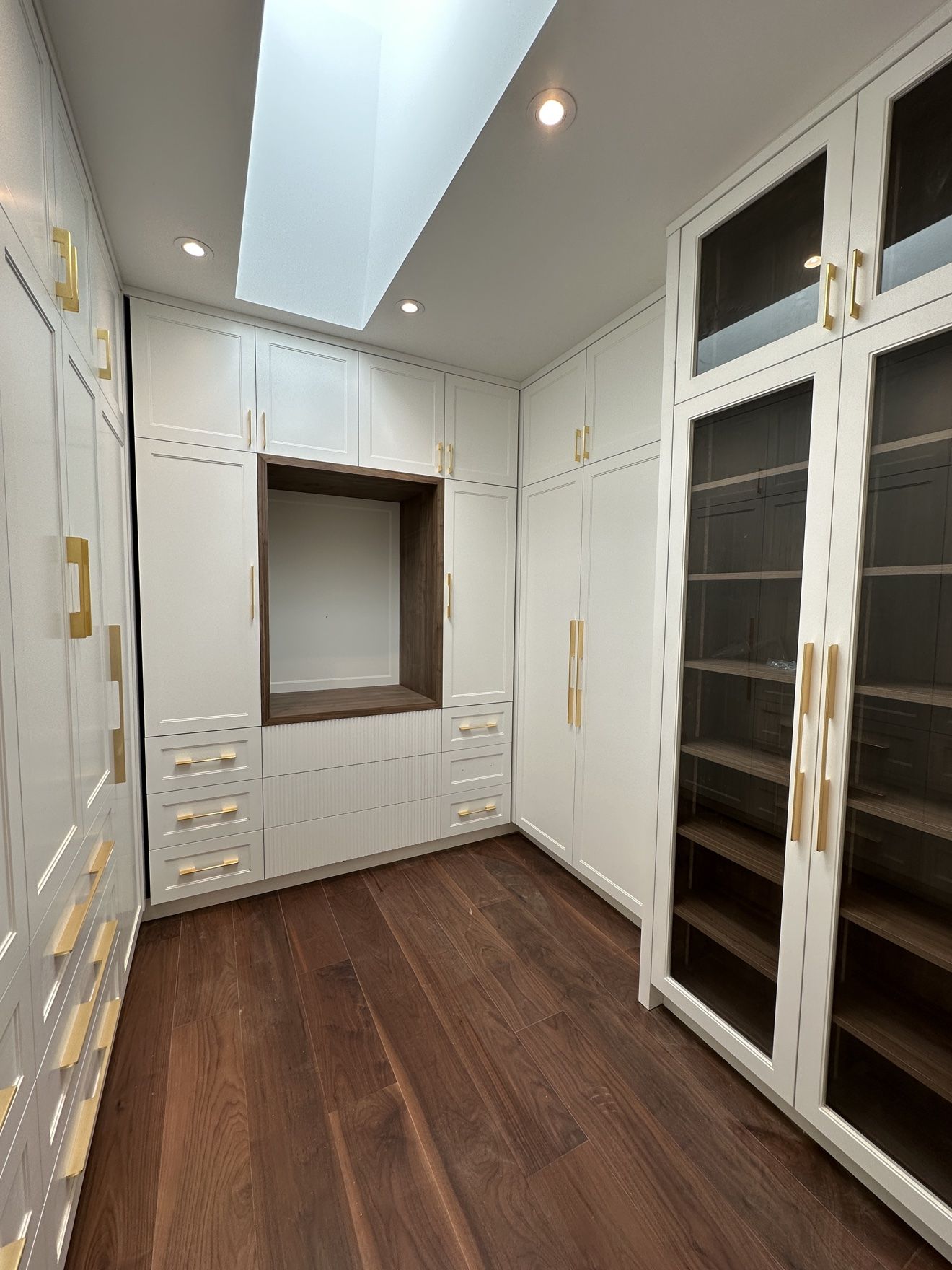 White walk-in closet with brass hardware, glass cabinets and skylight